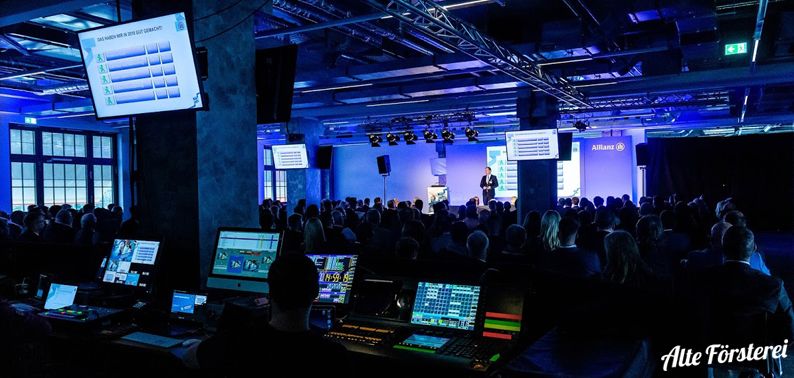 Konferenzraum mit Publikum, Bühne mit Redner und Projektionsbildschirmen, technischer Bereich mit Mischpulten im Vordergrund.