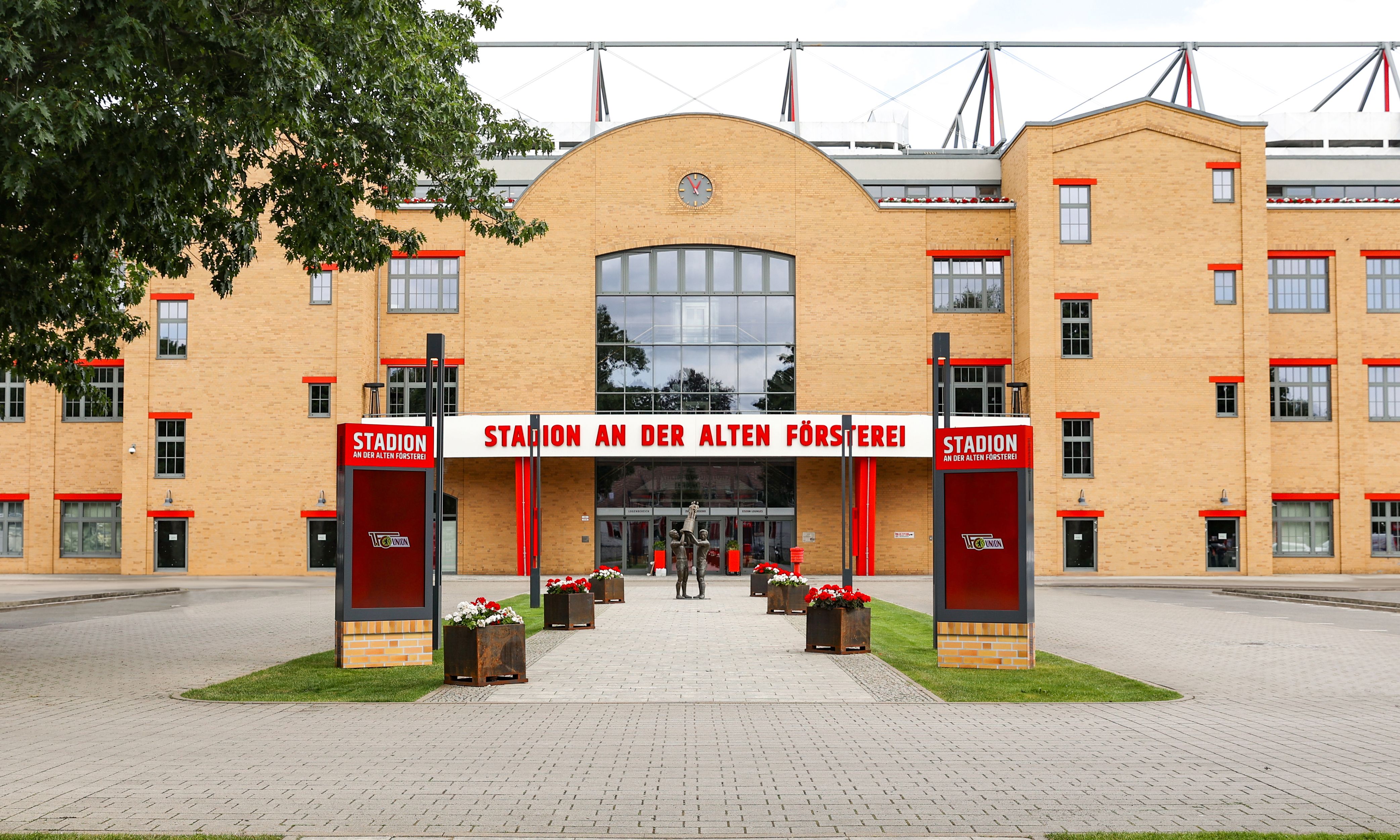 Stadion an der Alten Försterei mit rotem und weißem Gebäude, Blumenbeeten und Pforte auf einem gepflasterten Platz.