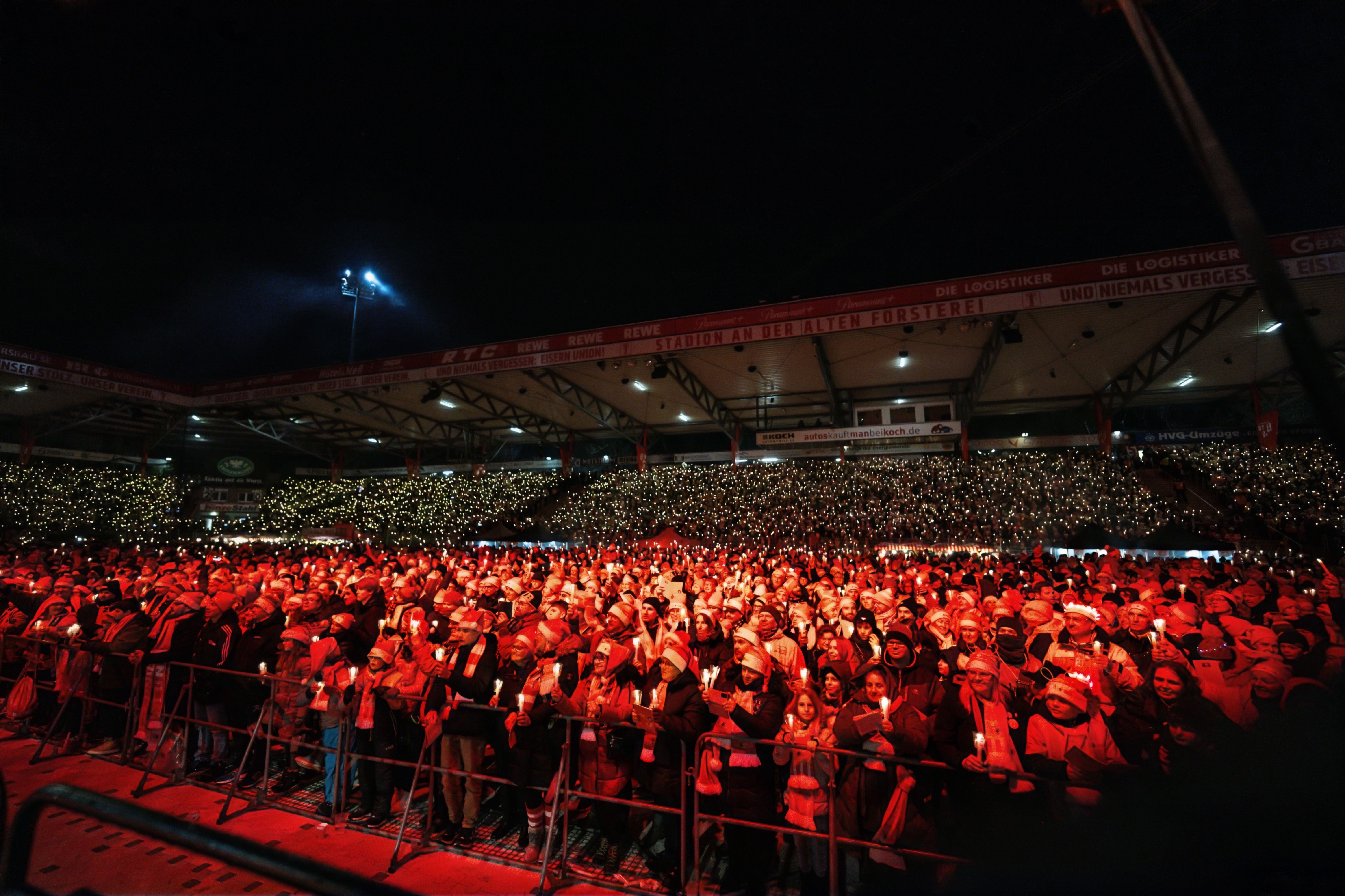 Ein großer Menschenmenge bei einem Konzert oder einer Veranstaltung, umgeben von bunten Lichtern und einer dunklen Nacht.
