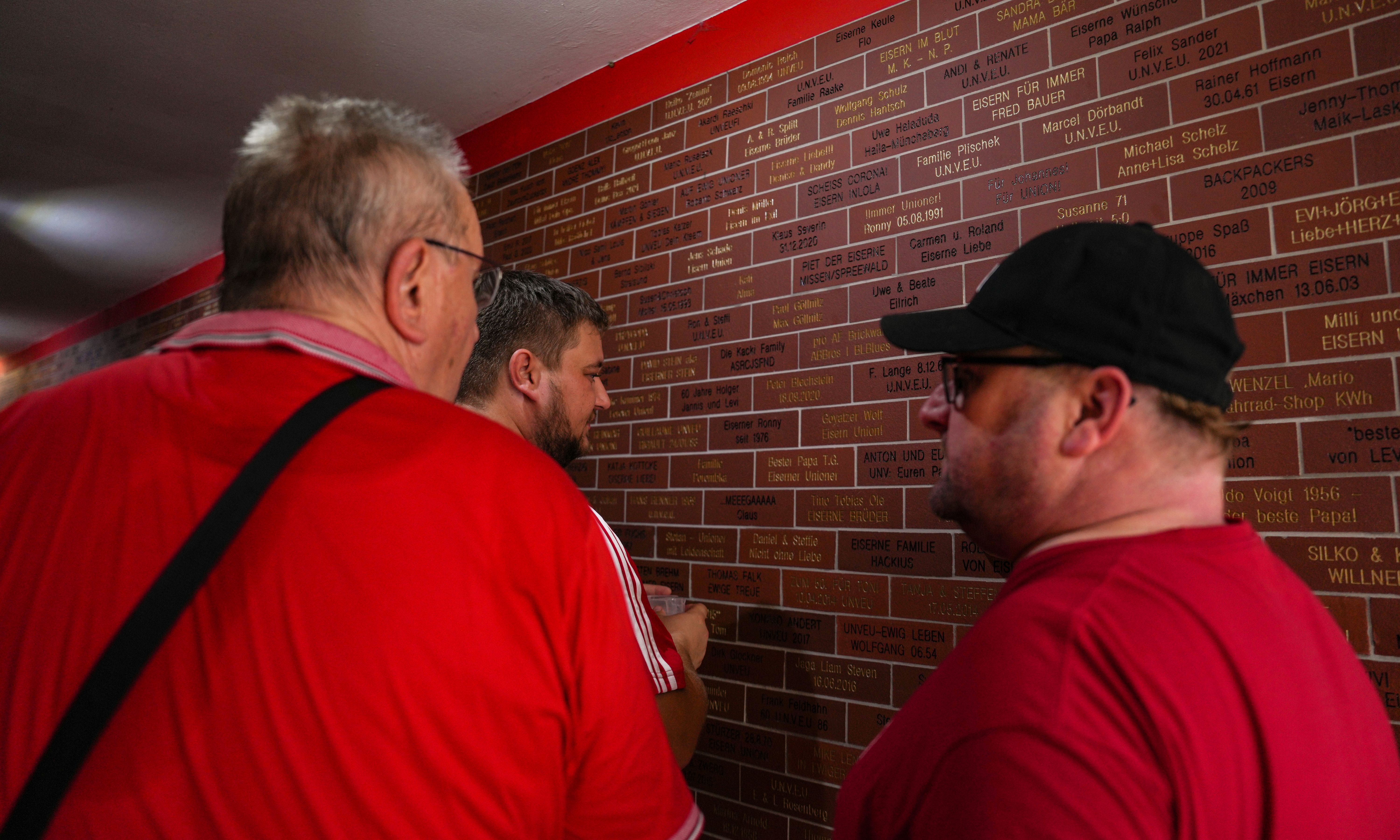 Drei Männer in roten Shirts betrachten eine mit Namen und Daten beschriftete Wand in einem Innenraum.